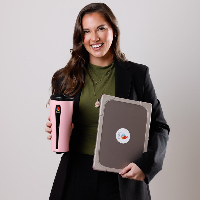 Sidney in a black blazer and green top holds a pink travel mug and a gray laptop against a light gray studio background.